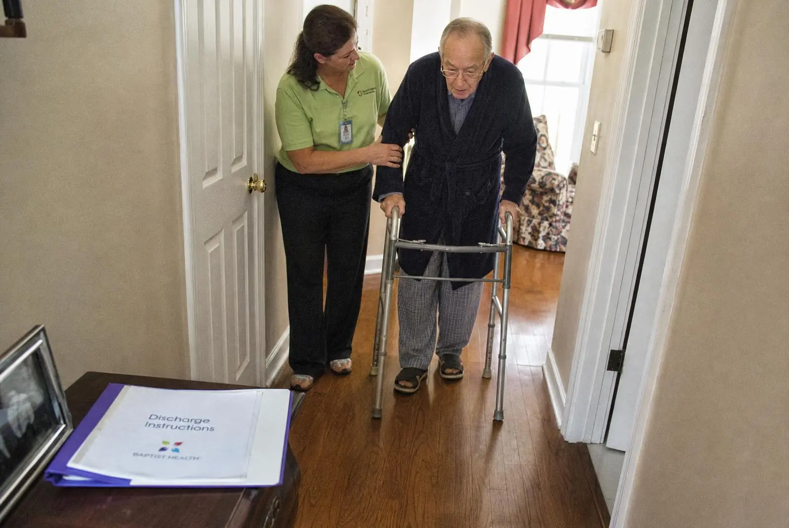 A caregiver steadying an older man as he walks through his warm, sunlit home